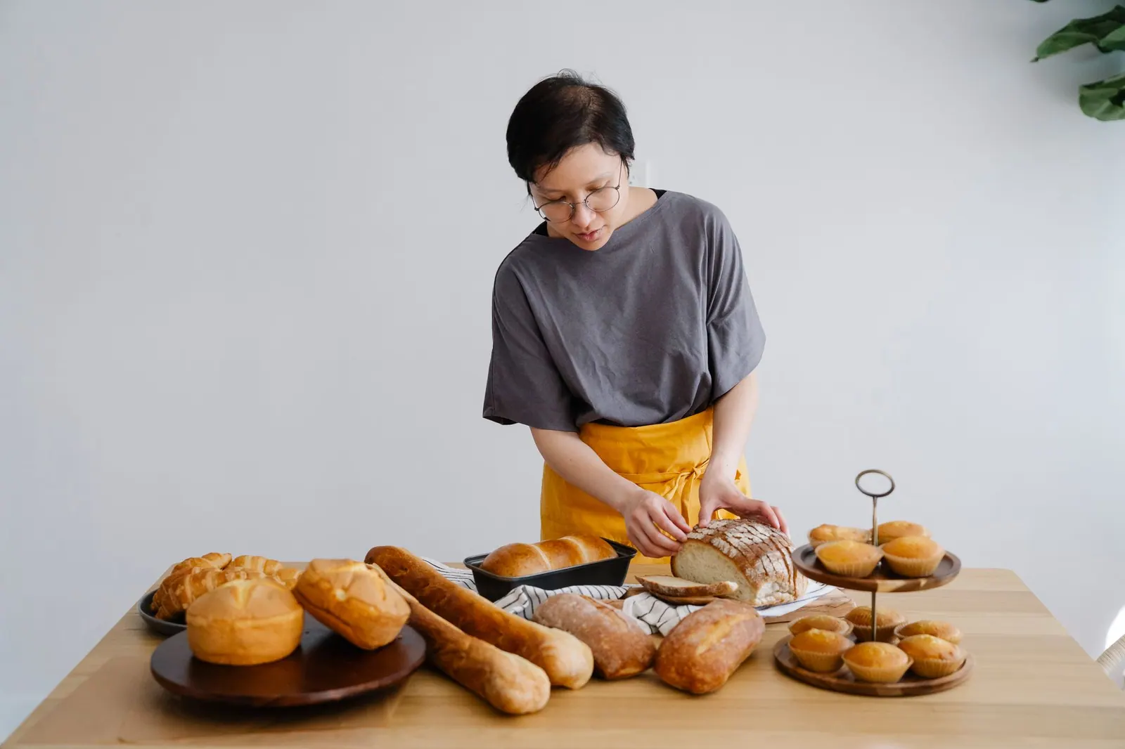 A baker in a yellow apron slices a fresh loaf at a table of breads and muffins